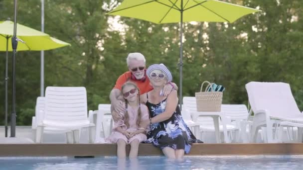 Retrato pareja madura abrazando a la nieta en el borde de la piscina. Abuela, abuelo y nieto saludando con las manos. Feliz familia amistosa. Descanse en el hotel. Recreación y ocio al aire libre — Vídeos de Stock