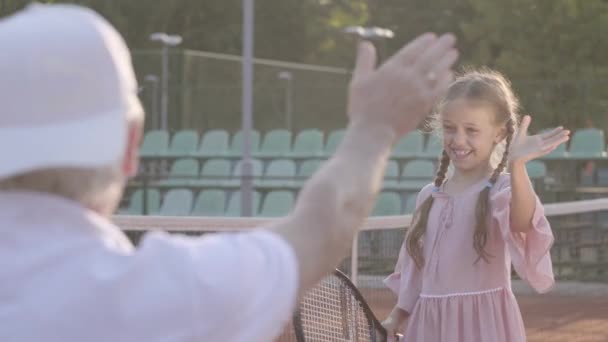 Sevimli küçük gülümseyen mutlu kız elinde bir tenis raket ile tenis kortu kameraya bakarak ayakta. Dedesi başparmak gösteren torunu sevinirler. — Stok video