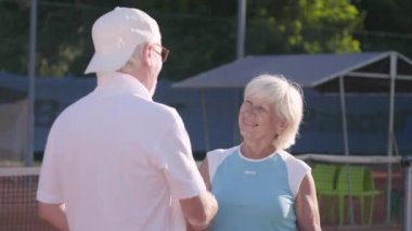 Gülümseyen olgun çift tenis kortu tenis oynadıktan sonra el sıkışıyor. Açık havada aktif boş zaman. Kıdemli erkek ve kadın birlikte eğleniyor