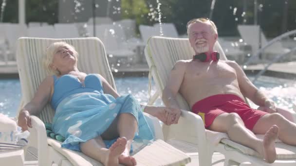 Pareja de ancianos positivos tumbados en tumbonas cerca de la piscina tomados de la mano y sonriendo. Feliz familia amorosa. Descanse en el hotel. Vista frontal — Vídeos de Stock