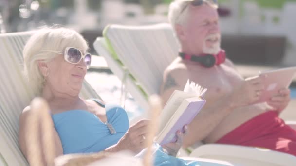 Schattig paar liggend op ligbedden bij het zwembad. Rijpe vrouw het lezen van een boek terwijl oude man kijken naar een Tablet. Gelukkig liefdevolle familie. Rust in Hotel. — Stockvideo