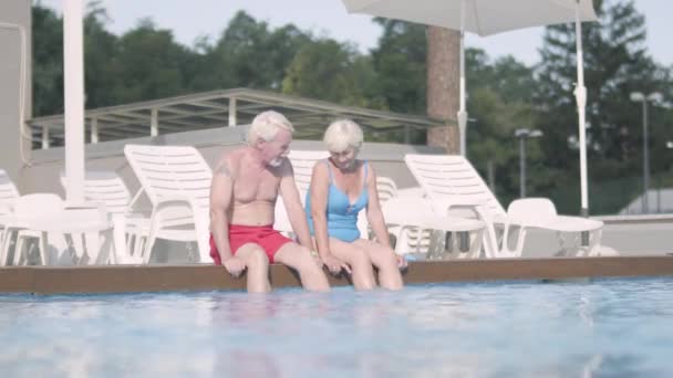 Glückliches älteres Paar, das am Rande des Pools sitzt. Netter älterer Herr und Frau, die sich entspannt in der Hotelanlage umarmen. glückliche, freundliche Familie. — Stockvideo