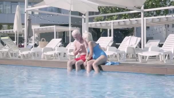 快乐成熟的夫妇坐在池边，他们的脚在水中。可爱的老年男人和女人在酒店大楼里放松。幸福友好的家庭。酒店休息 — 图库视频影像
