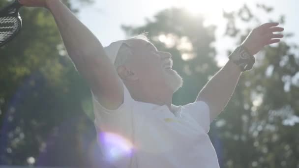 Успешный позитивный улыбающийся зрелый мужчина играет в теннис на теннисном корте. Старик бросает мяч ракеткой. Активный отдых на свежем воздухе. Медленное движение . — стоковое видео