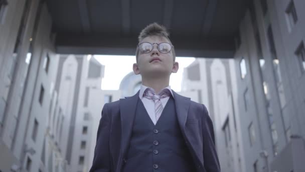 Ritratto carino ragazzo ben vestito in piedi in vista sulla strada guardando in macchina fotografica. Bambino serio di fronte a un bellissimo paesaggio urbano con edifici alti, grattacieli. Piccolo abitante della citta '. Bambino come — Video Stock