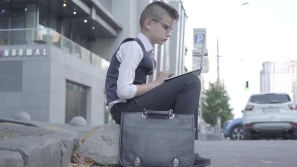 Entzückender, gut gekleideter Junge mit Brille, der auf der Straße sitzt und in Großaufnahme am Laptop arbeitet. ernstes Kind, das sich wie ein Erwachsener verhält. Der Junge ist geschäftlich tätig. Kind als Erwachsener. — Stockvideo