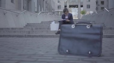 Yakışıklı iyi giyimli çocuk sokakta merdivenlerde oturan arka planda ev işi yapıyor. Ön planda asfalt üzerinde duran eski çanta. Kamera sağa ve sola hareket ediyor
