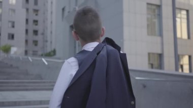 Omzunda ceketi olan yakışıklı ve iyi giyimli bir çocuğun arka görünümü sokakta durup binaya bakıyor. Gökdelenler ile güzel şehir manzarası önünde ciddi çocuk. Küçük şehir lisacısı.