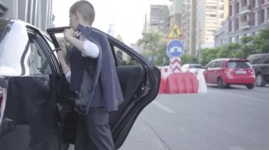 Ciddi iyi giyimli çocuk arabadan iniyor ve sokakta yürüyor. Arka planda Kentsel şehir manzarası. Zengin babanın küçük çocuğu. Yetişkin olarak çocuk. Geri görünüm
