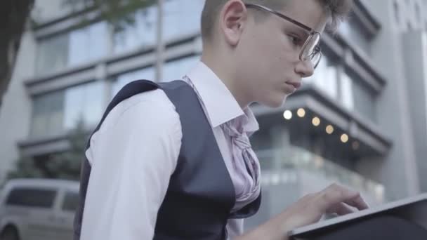 Snygg välklädd pojke i glasögon sitter på gatan arbetar på den bärbara datorn närbild. Allvarliga Kid agerar som vuxen. Pojken är engagerad i näringslivet. Barn som vuxna. — Stockvideo