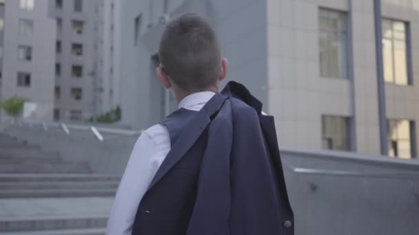 Achteraanzicht van een knappe goed geklede jongen met jas op zijn schouder staande op de straat kijken naar gebouw. Serieus kind voor een prachtig stadsbeeld met wolkenkrabbers. Little City Dweller. — Stockvideo