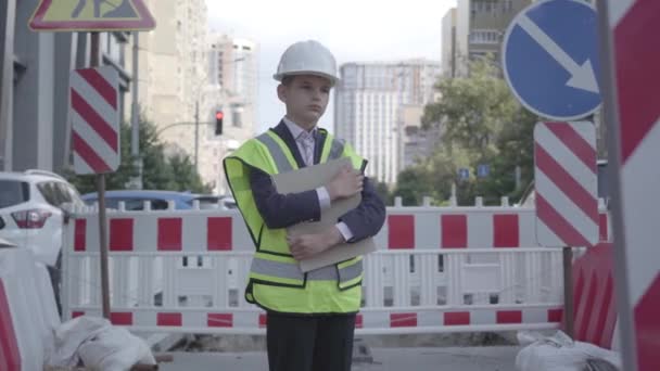 大都会の道路標識やピットを背景に、混雑した道路に立つ建物計画を保持する安全装置とコンストラクターヘルメットを身に着けている思慮深い小さな男の子。大人の子供. — ストック動画