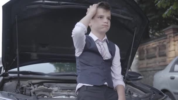Upset little boy in a business suit sitting next to the open hood of a broken car. Child as adult. — Stock Video