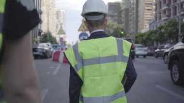 Küçük yol işçisi veya müfettiş güvenlik ekipmanları ve konstrüktör kask tanınmayan yardımcısı ile yürüyüş giyen büyük bir şehirde yoğun bir yolda acil bir siteye yürüyor. Yetişkin olarak çocuk.