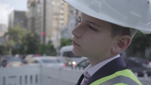 Lindo niño pensativo con traje de negocios y equipo de seguridad y casco de constructor de pie en una carretera concurrida en una gran ciudad. Ingeniero, arquitecto, constructor haciendo su trabajo. Niño como adulto — Vídeos de Stock