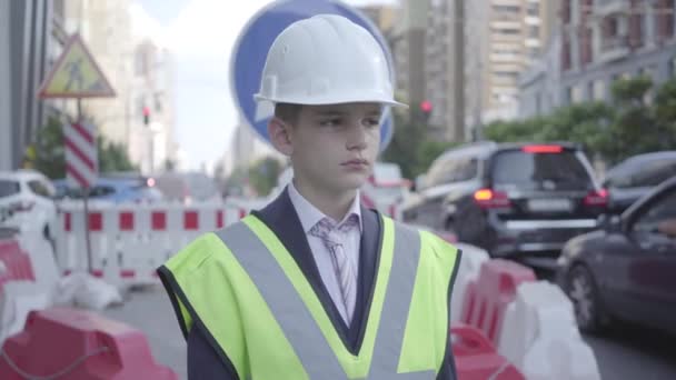 Portret schattige kleine jongen dragen Business Suit en veiligheidsuitrusting en constructeur helm staande op een drukke weg in een grote stad. Ingenieur, architect, bouwer doet zijn werk. Kind als volwassene. — Stockvideo