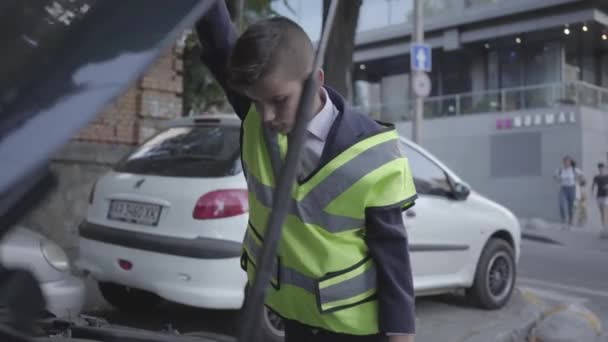 Sebevědomý chlapec, který nosí bezpečnostní vybavení stojící u otevřeného kápě rozbitého auta. Ten kluk opravuje auto. Dítě jako dospělý. — Stock video