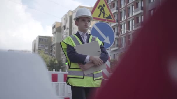 Portret doordachte kleine jongen dragen veiligheidsuitrusting en constructeur helm houden bouwplan staande op een drukke weg op de achtergrond van verkeersborden in een grote stad. Kind als volwassene. — Stockvideo