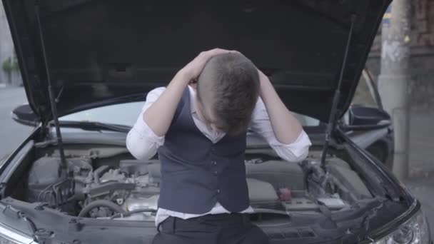 Le jeune garçon stressé bien habillé debout devant la voiture cassée avec un capot ouvert. Le garçon fait l'expérience du résultat de ses devinettes — Video