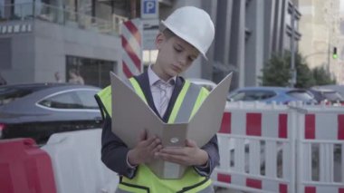 Portre küçük çocuk güvenlik ekipmanları ve inşaat kask okuma bina planı giyen, büyük bir şehirde yol işaretleri arka planda yoğun bir yolda iş kontrol. Yetişkin olarak çocuk.