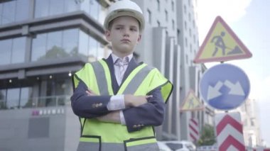 Portre sevimli küçük başarılı çocuk iş elbisesi ve güvenlik ekipmanları ve inşaat kask büyük bir şehirde yoğun bir yolda ayakta giyiyor. Mühendis, mimar, inşaatçı işini yapıyor. Yetişkin olarak çocuk