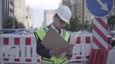 Portre küçük çocuk güvenlik ekipmanları ve inşaat kask okuma bina planı giyen, büyük bir şehirde yol işaretleri arka planda yoğun bir yolda iş kontrol. Yetişkin olarak çocuk.