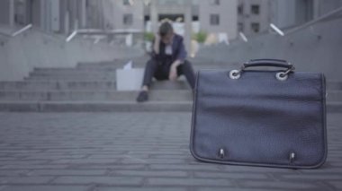 Yakışıklı küçük çocuk arka planda ev işleri yapıyor sokakta merdivenlerde oturan. Ön planda asfalt üzerinde duran eski çanta. Kamera sağa ve sola hareket ediyor