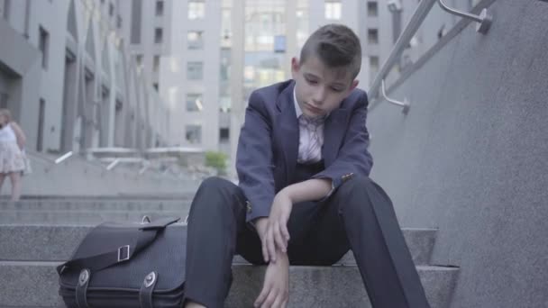 Pensive schattige jongen dragen Business Suit zittend op de trap op de straat. De jongen is moe en wil gewoon een kind zijn. Kind als volwassene. — Stockvideo