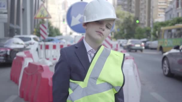 疲惫的小男孩头戴建筑头盔，统一看着摄像机，竖起大拇指。架构师概念。工程师，建筑师，建筑工人做他的工作。儿童成人 — 图库视频影像