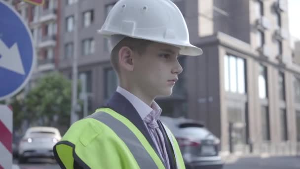 Портрет симпатичного мальчика в деловом костюме и защитном снаряжении и шлеме конструктора, стоящего на оживленной дороге на фоне дорожных знаков в большом городе. Ребенок как взрослый . — стоковое видео