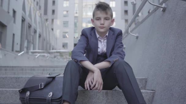 通りの階段に座っているペンシブな服を着たかわいい男の子。その少年は疲れていて、ただの子供になりたがっている。大人としての子供. — ストック動画