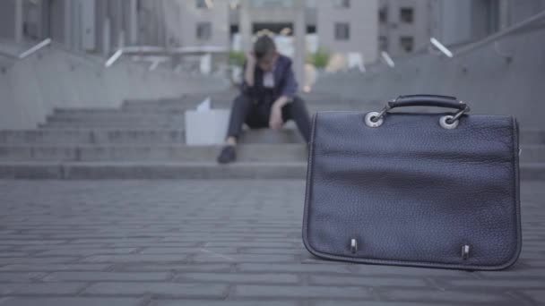 Handsome small boy sitting on the stairs on the street doing home work at the background. Old purse standing on the asphalt in the foreground. Camera moving right and left — Stock Video