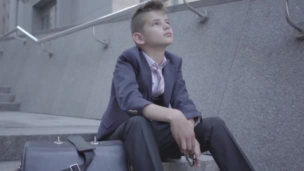 Triste chico bien vestido sentado en las escaleras de la calle. El chico está cansado y quiere ser solo un niño. Niño como adulto . — Vídeo de stock
