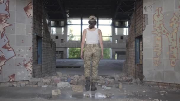 Joven confiada en uniforme militar caminando lentamente en polvoriento edificio sucio abandonado. Mujer guerrera en una fábrica desierta. Mujer soldado — Vídeo de stock