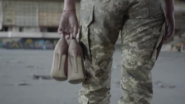 Piernas de mujer joven en uniforme militar caminando lentamente en polvoriento edificio abandonado sucio llevando zapatos de tacón alto en las manos. El concepto de una chica fuerte y femenina. Mujer soldado — Vídeos de Stock