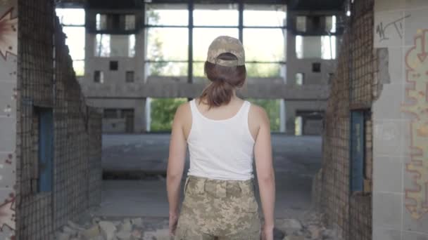 Atractiva joven con uniforme militar caminando lentamente en un sucio y polvoriento edificio abandonado. Mujer guerrera en una fábrica desierta. Mujer soldado — Vídeos de Stock