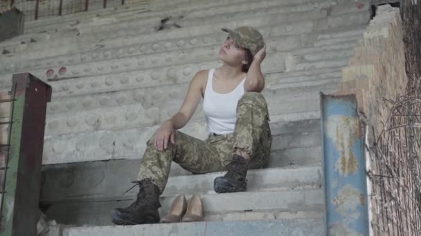 Giovane donna triste in uniforme militare seduta sulle fredde scale di cemento nell'edificio abbandonato. Scarpe col tacco alto vicino. Donna soldato — Video Stock