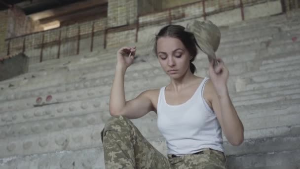 Красивая молодая женщина в военной форме сидит на холодной бетонной лестнице в заброшенном здании. Девушка растворяет волосы, демонстрируя свою женственность. Концепция сильной, но женственной девушки — стоковое видео