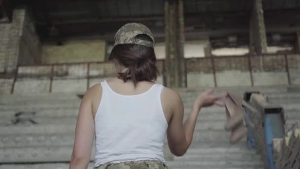 Une jeune femme en uniforme militaire mettant ses chaussures à talons hauts sur son épaule et regardant dans la caméra souriante. Le concept d'une femme forte. Vue de derrière. Femme soldat — Video