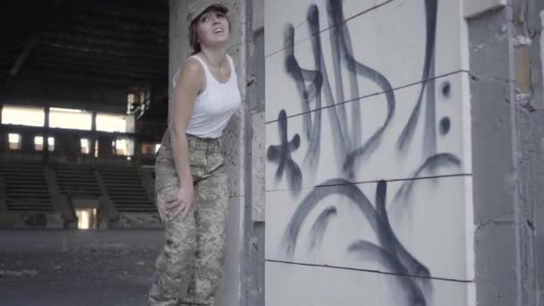 Giovane donna in uniforme militare che si nasconde dal nemico in un edificio abbandonato sporco e polveroso. Ragazza magra correndo verso la fotocamera e fermandosi a riprendere fiato. Il concetto di azioni militari, salvare vite umane — Video Stock