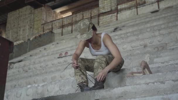 Mladá žena ve vojenské uniformě, která sedí na chladném betonových schodech v opuštěné budově a rozvazuje si boty, aby se připravovala na trénink. Boty na vysokém podpatku. Žena bojovnice v — Stock video