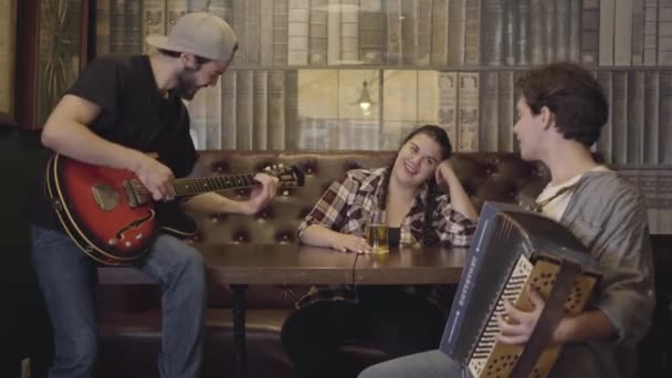 Joven hombre barbudo sonriente tocando la guitarra en el bar, su amigo tocando el acordeón mientras atractiva mujer regordeta sentada en la mesa bebiendo cerveza. Ocio en el pub. Amigos divirtiéndose juntos — Vídeo de stock