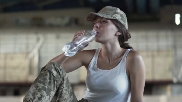 Mulher jovem bonito em uniforme militar água potável da garrafa sentado no chão em edifício abandonado sujo empoeirado. Mulher guerreira numa fábrica deserta. Mulher soldado — Vídeo de Stock