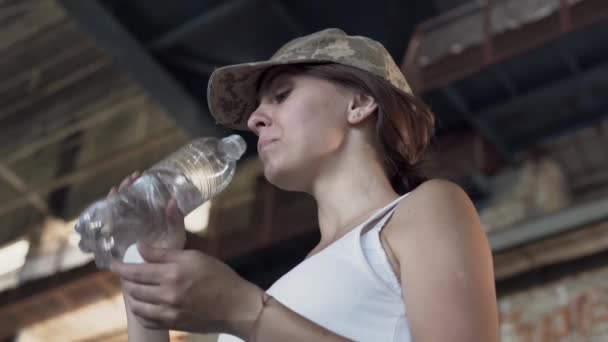 Portrait of pretty young woman in military cap drinking water from the bottle in dusty dirty abandoned building. The concept of strong but feminine girl. Human life in wartime. Bottom view — Stock Video