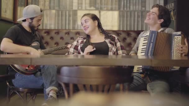 Joli jeune homme barbu souriant jouant de la guitare dans le bar, son ami jouant de l'accordéon tandis qu'adorable femme dodue assise près de sourire. Loisirs au pub. Amis s'amuser ensemble — Video