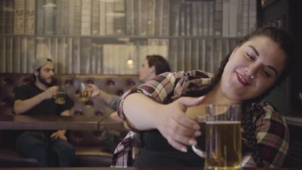 Mujer regordeta con coletas sentada en el mostrador del bar con un vaso de cerveza mientras dos hombres hablan con entusiasmo bebiendo alcohol en el fondo. Ocio en el bar — Vídeo de stock
