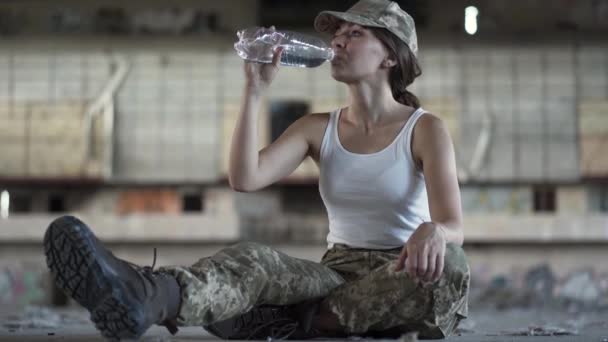 Atractiva joven con uniforme militar bebiendo agua de la botella sentada en el suelo en un sucio y polvoriento edificio abandonado. Mujer guerrera en una fábrica desierta. Mujer soldado — Vídeos de Stock