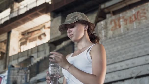 ほこりっぽい汚れた放棄された建物のボトルから水を飲む軍帽の魅力的な若い女性の肖像画。無人工場の戦士の女性女性兵士 — ストック動画