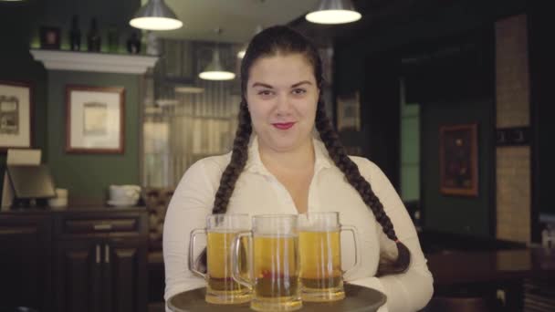 白いブラウスにピグテールを着たふっくらした女性は、カメラに微笑む3つのビールグラスを持つトレイを持っています。男性の手は、トレイのクリンキングメガネからアルコールの2つのグラスを取ります。バーでのレジャー — ストック動画