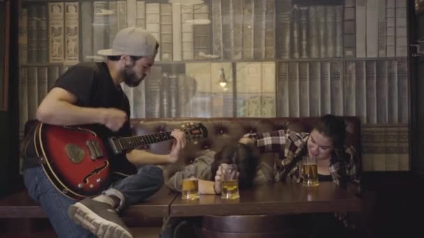 Jeune homme barbu positif jouant de la guitare dans le bar, ses amis assis près. Loisirs au pub. Les gars et la fille s'amusent ensemble . — Video